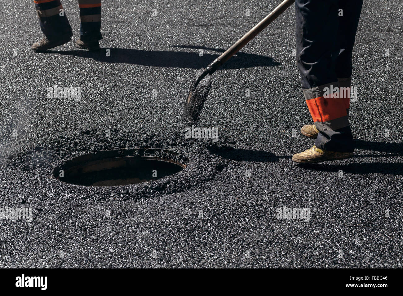 En construction routière en milieu urbain, l'asphaltage en cours, travailleur avec pelle près d'égout, fragment de pieds Banque D'Images