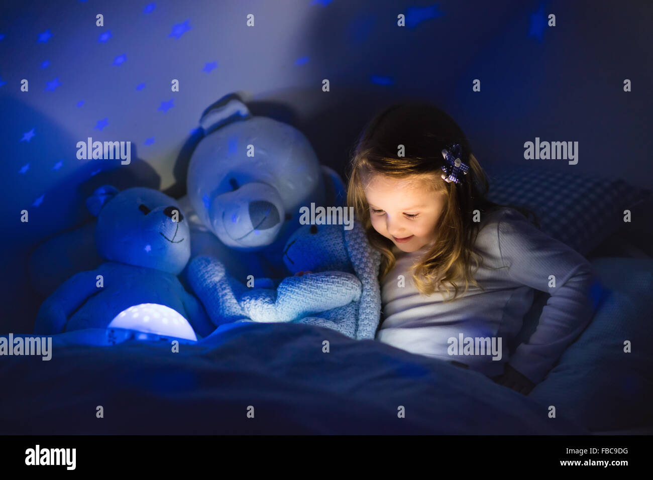 Petite fille lire un livre au lit. Chambre sombre avec la projection de lumière nuit étoile sur le prix plafond. Les enfants et la literie de pépinière. Banque D'Images
