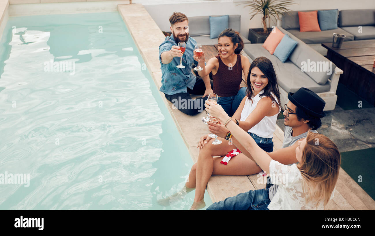 High angle shot de jeunes gens assis au bord de la piscine et du vin d'avoir le sourire. Groupe de société multiraciale friends toasting at pool party Banque D'Images