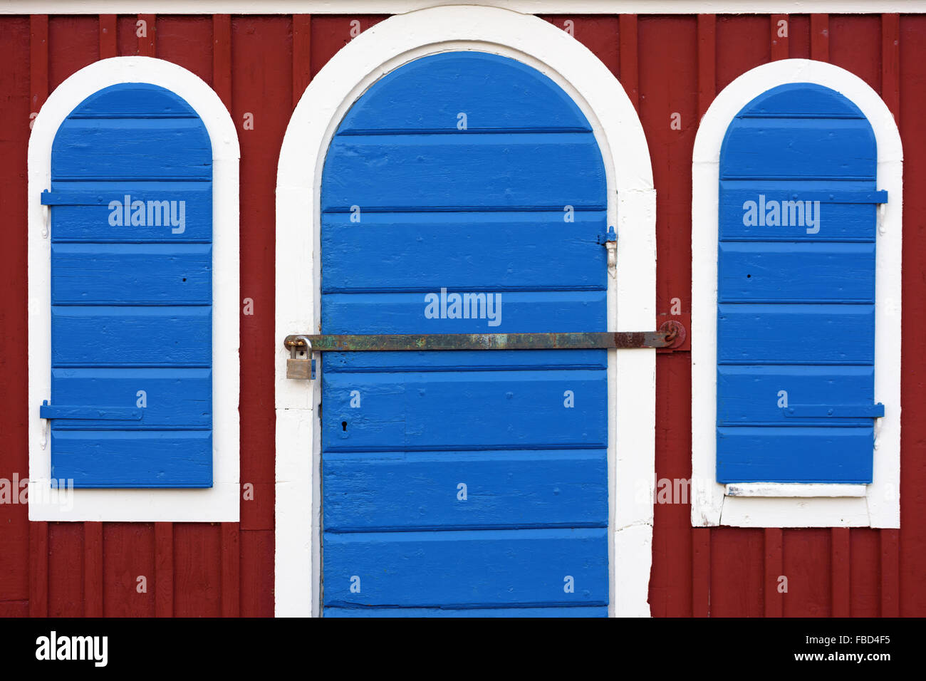 Porte et volets bleus sur une cabane rouge. Porte et fenêtres sont encadrées de blanc. Barre de fer devant la porte est verrouillée par un cadenas. Banque D'Images