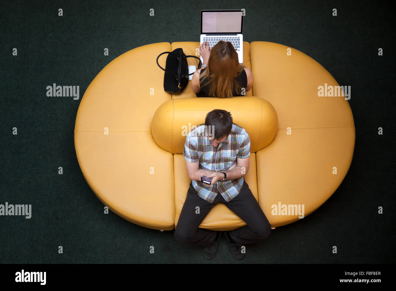 Les jeunes, hommes et femmes, assis sur yellow coach dos à dos, à l'aide de dispositifs électroniques dans la zone wifi public, travaillant sur la Banque D'Images