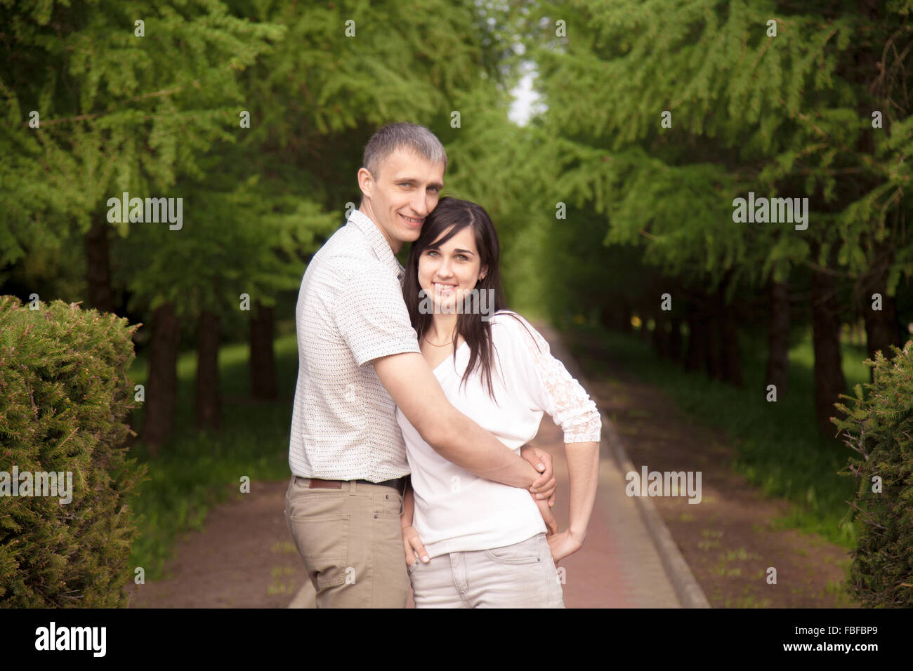 Portrait of smiling couple, jeune homme et brunette woman standing on walkway dans parc, enlacés Banque D'Images