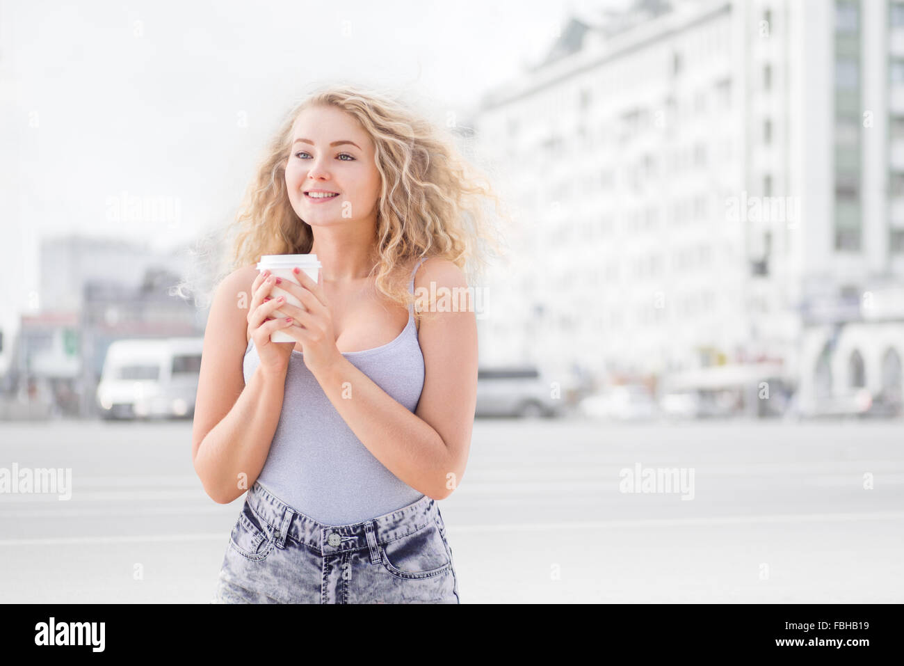 Heureux jeune femme avec de longs cheveux bouclés, tenant une tasse de café à emporter et souriant avec flirter devant une caméra contre les Banque D'Images