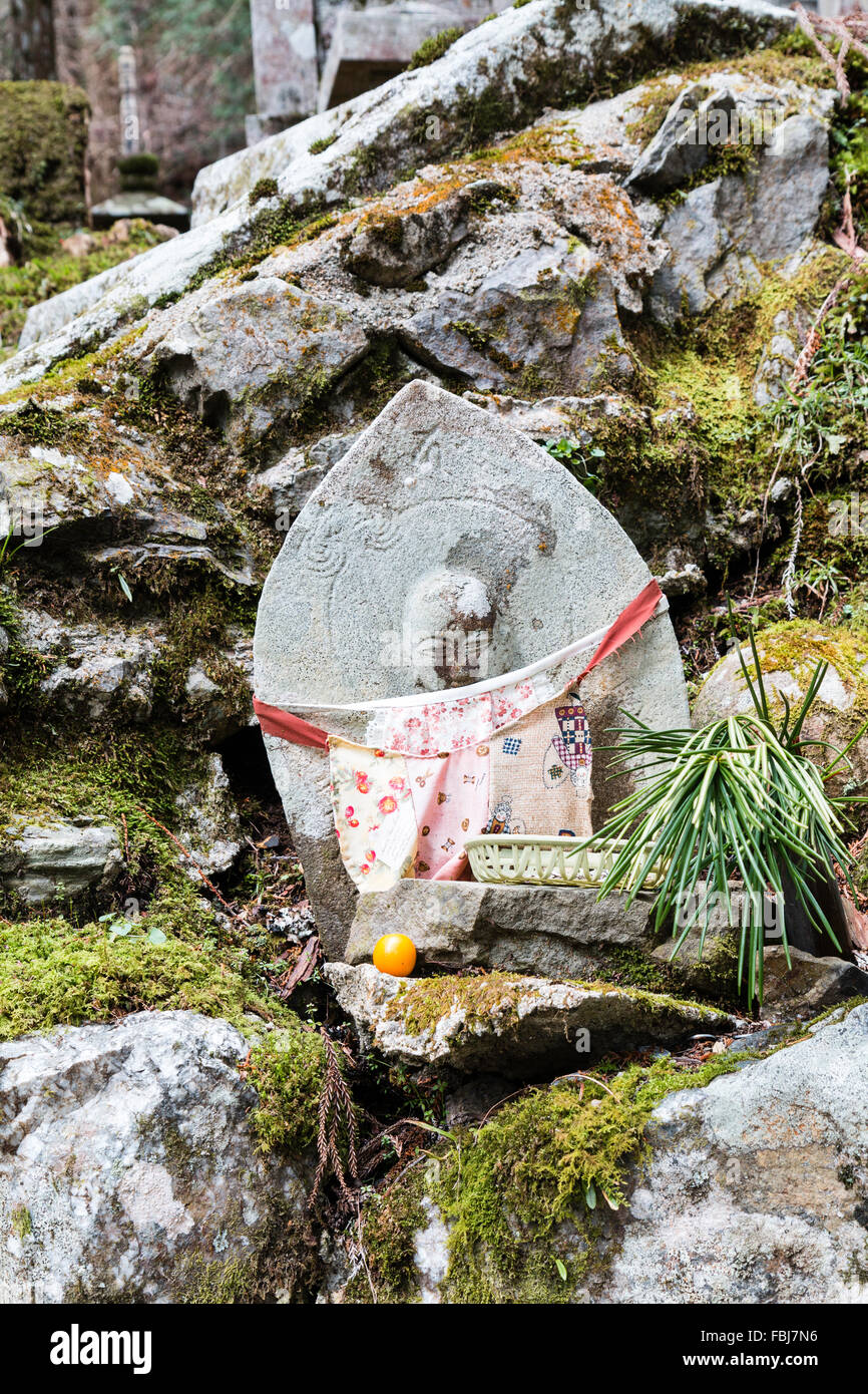 Koyasan, Okunoin cemetery. Petit rouge pâle bibbed statue Jizo Bosatsu, 'Stone Bouddha', avec une offre de fruits orange, debout sur des rochers. Banque D'Images