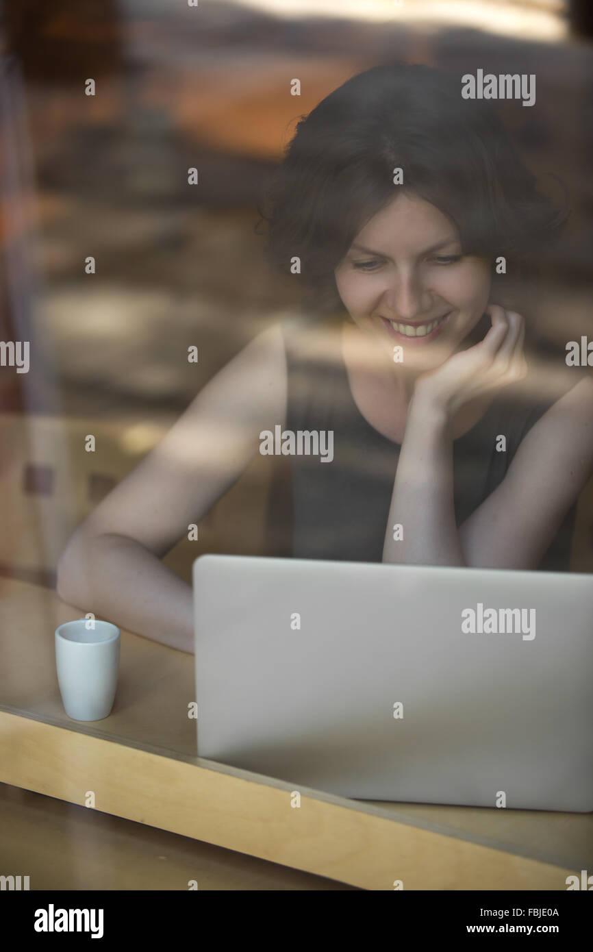 Portrait of happy smiling beautiful young woman sitting in cafe moderne avec une tasse de boisson sur la table, using laptop Banque D'Images