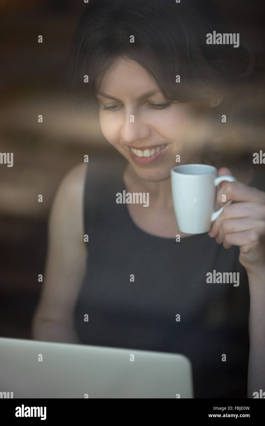 Portrait of beautiful happy smiling young woman sitting in cafe moderne holding tasse de café, working on laptop Banque D'Images