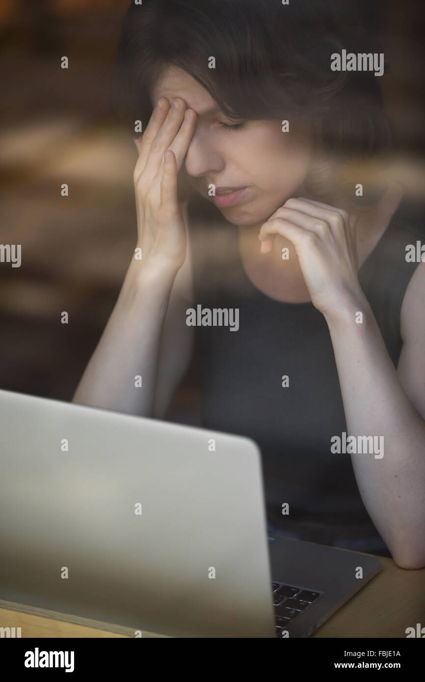 Jeune femme assise dans le café en face de l'ordinateur portable, appuyé sur sa main, toucher la tête avec l'expression fatiguée, avoir des maux de tête Banque D'Images