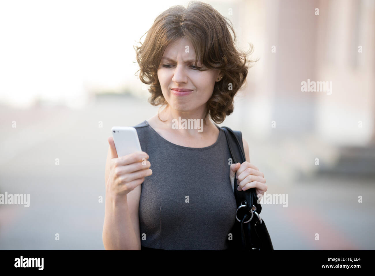 Portrait of young woman holding a souligné office téléphone cellulaire dans les mains sur la rue de la ville en été, en regardant l'écran avec cross Banque D'Images