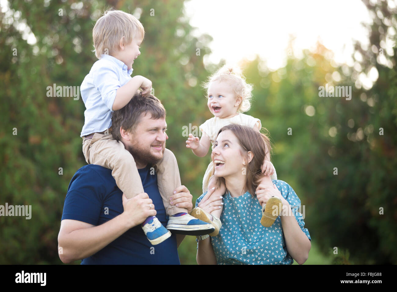 Portrait of happy family of four walking in park en été. Maman et papa transportant deux petits enfants sur rire joyeux Banque D'Images