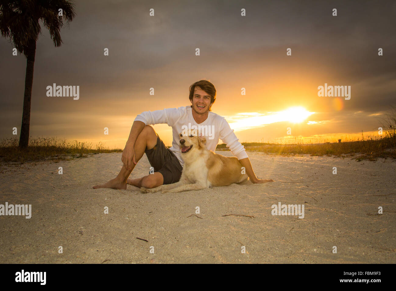 Homme assis sur plage avec golden retriever dog au coucher du soleil Banque D'Images