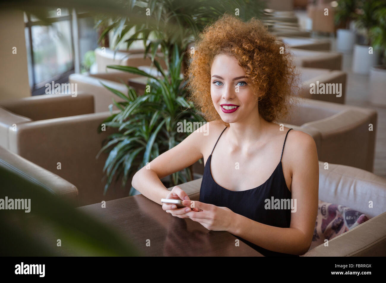 Heureuse femme rousse avec les cheveux bouclés à la table et l'utilisation de smartphone dans restaurant Banque D'Images