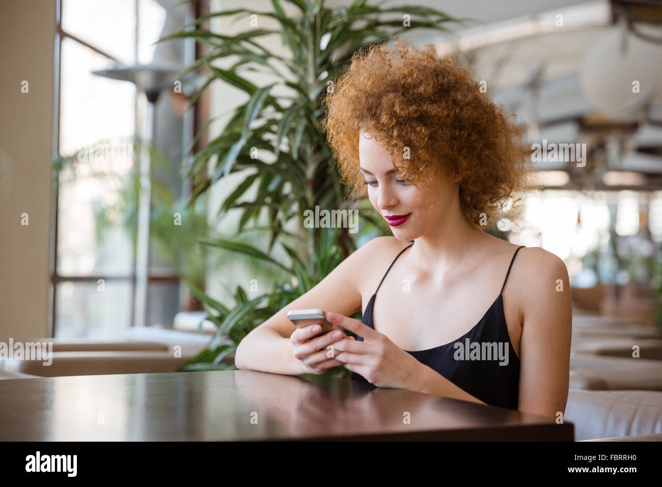 Femme rousse avec les cheveux bouclés à la table et l'utilisation de smartphone dans restaurant Banque D'Images