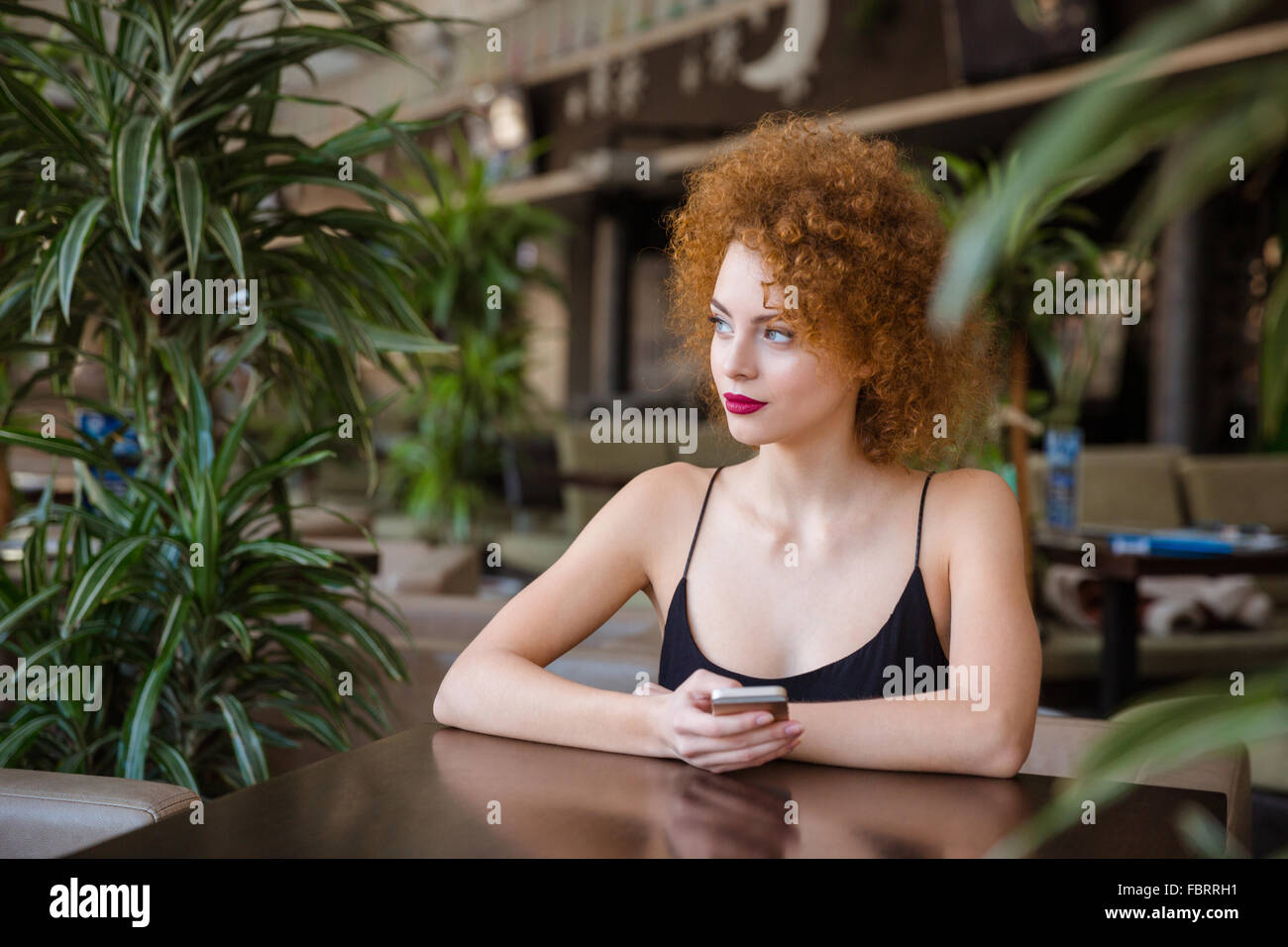 Thoughtful redhead femme avec les cheveux bouclés à la table avec le smartphone en restaurant et à l'écart Banque D'Images