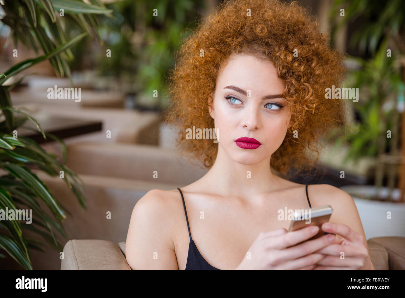 Femme rousse pensive avec des cheveux bouclés assis à la table avec le smartphone en restaurant et à l'écart Banque D'Images