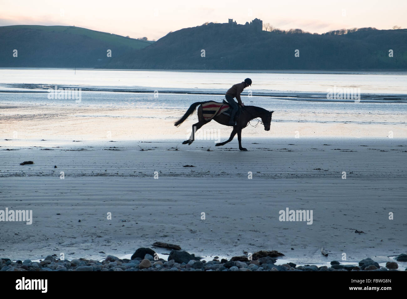 Ferryside, Carmarthenshire, Pays de Galles, Royaume-Uni. 19 janvier, 2016. Après des jours de pluie sur une journée ensoleillée au coucher du soleil/coucher du soleil.Si amèrement froid un jockey son cheval galope sur la plage vide à Ferryside, Carmarthenshire, Pays de Galles, U.K.Llanstephan Castle en arrière-plan. Crédit : Paul Quayle/Alamy Live News Banque D'Images