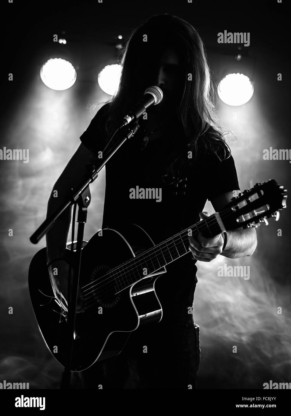 Photo d'un jeune homme en contre-jour avec des cheveux longs en silhouette jouant une guitare acoustique sur scène. Banque D'Images