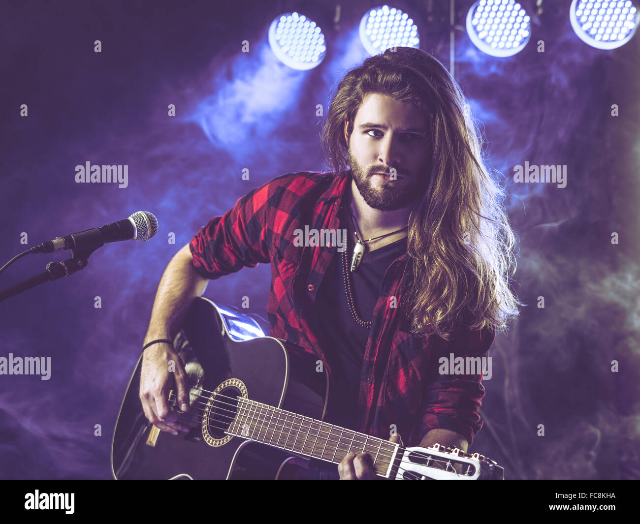 Photo d'un jeune homme aux cheveux longs et une barbe jouant une guitare acoustique sur scène avec des lumières et les concerts atmosphère. Banque D'Images