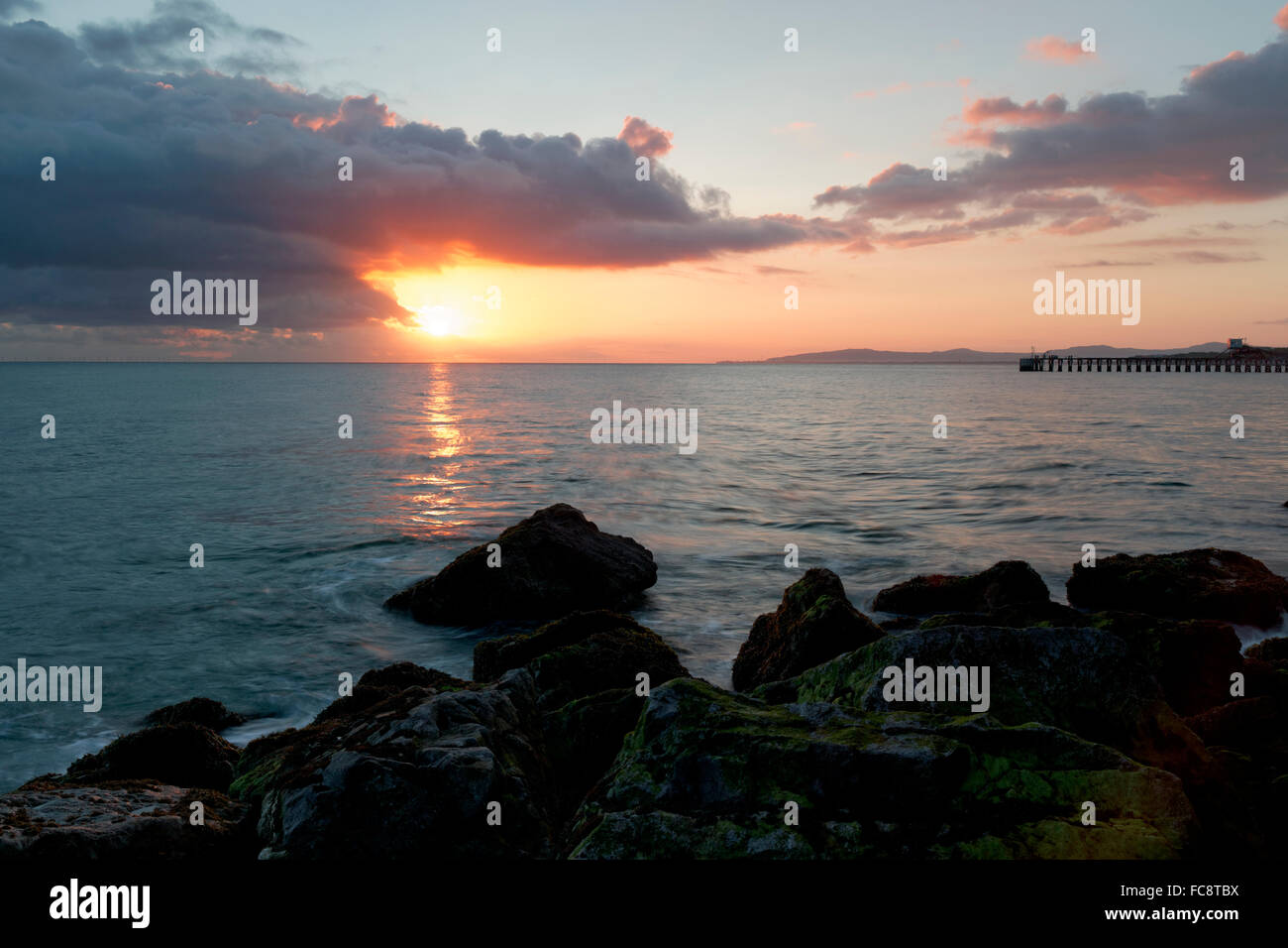 Paysage, Paysage marin, lever du soleil à travers les nuages au-dessus de la mer, jetée à distance, rochers en premier plan Banque D'Images