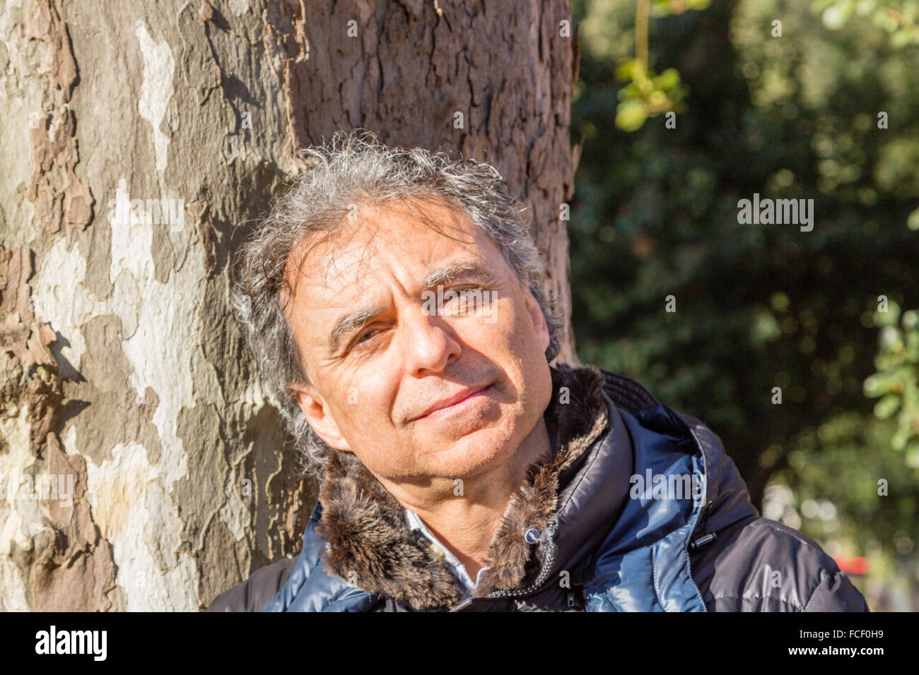 Homme de race blanche d'âge moyen aux cheveux gris vêtus de noir Down jacket appuyé contre un tronc d'arbre Banque D'Images
