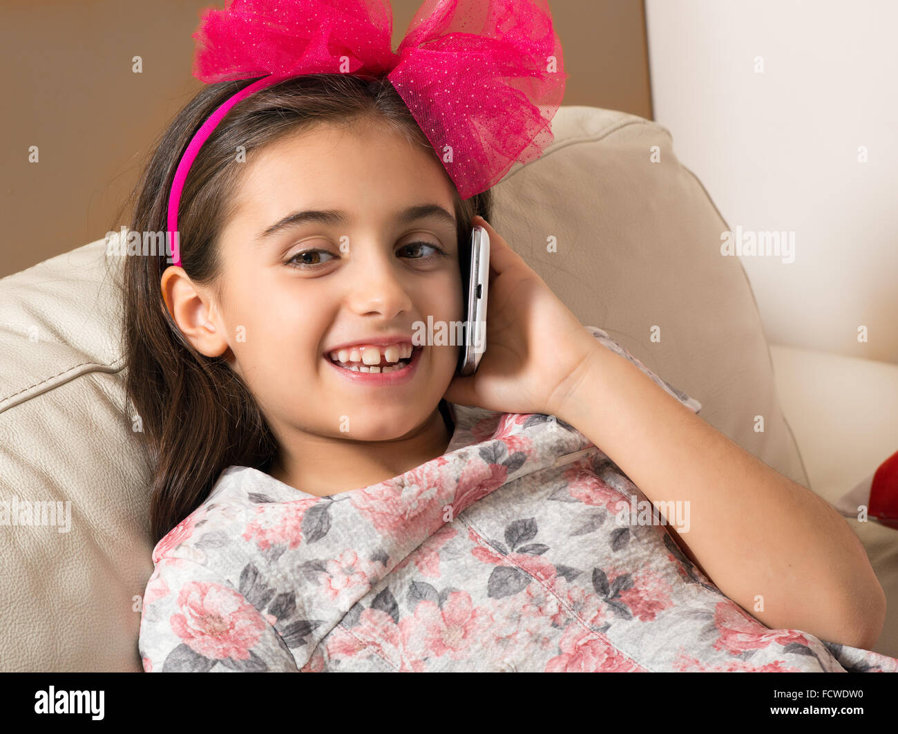 Jolie petite fille avec un joli sourire heureux et d'un noeud rose dans les cheveux de vous détendre sur un canapé chat sur son téléphone portable, Banque D'Images