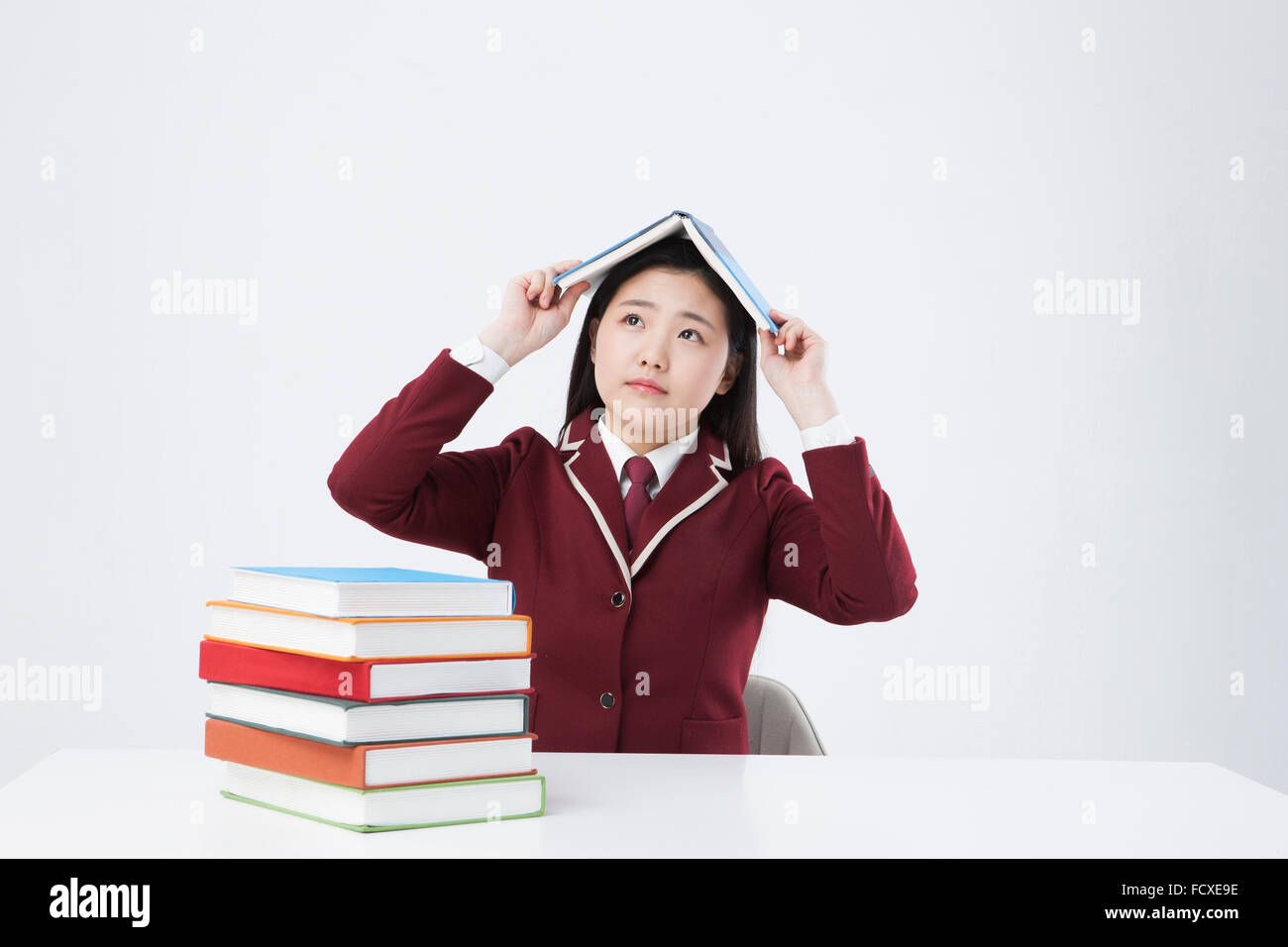 High school girl assis au bureau avec une pile de livres tenant un livre ouvert sur sa tête, et à la recherche jusqu'au visage inquiet Banque D'Images