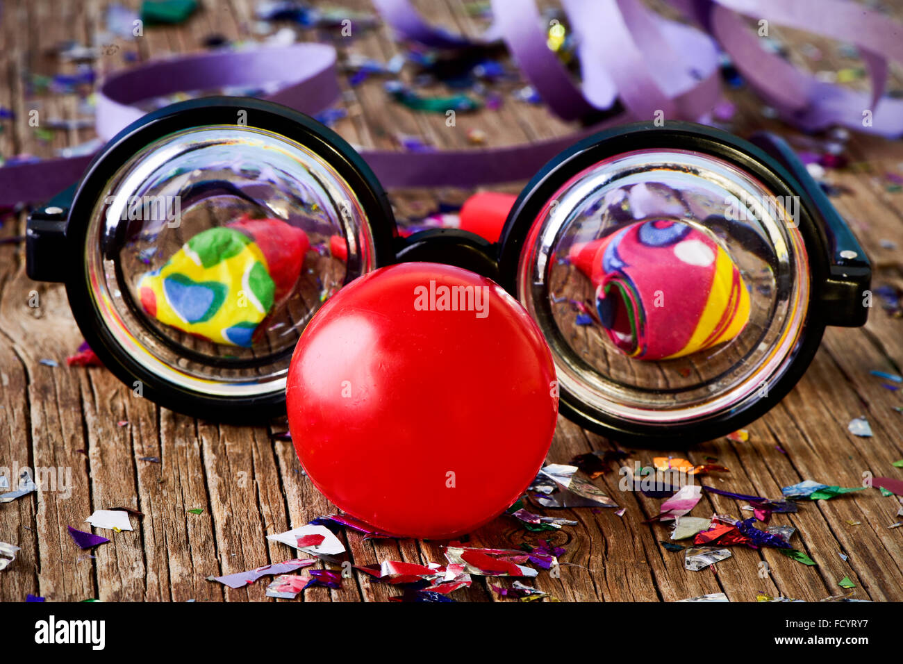 Une paire de fausses lunettes à courte vue et un nez de clown rouge, sur une surface en bois rustique plein de confettis, cornes et parti strea Banque D'Images