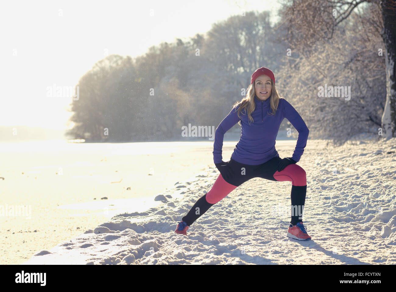 Jeune femme active l'exercice en plein air dans un parc à neige de l'hiver blanc frais faisant des exercices d'étirement pour réchauffer comme elle sourit Banque D'Images