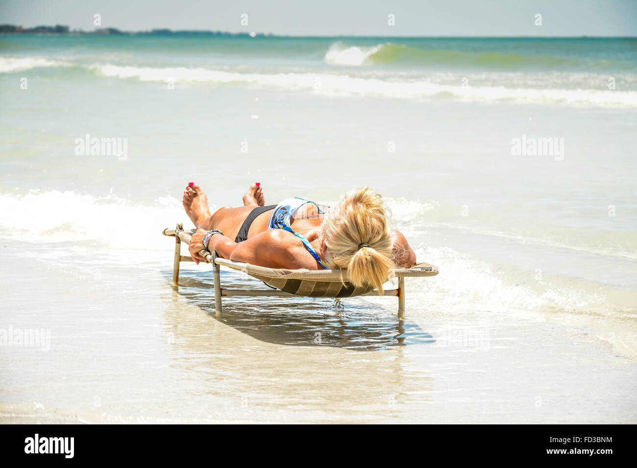 Une femme solo se trouve face vers le haut et à l'horizontale sur une chaise longue plage entourée par le fracas des vagues doucement du surf à la plage Banque D'Images