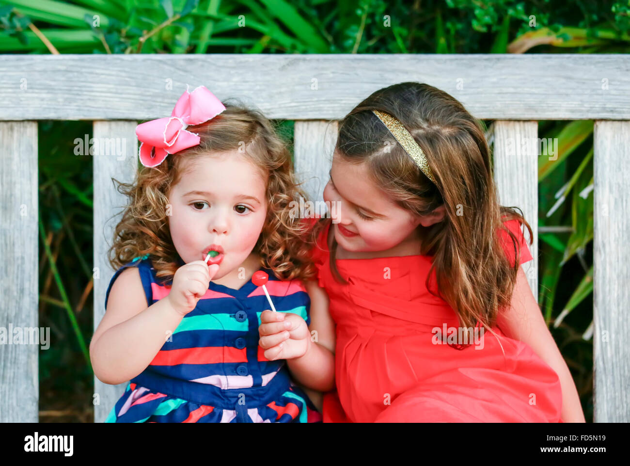 Deux petites filles en robes de couleur vive assis sur un banc, profitant de lollipops. Banque D'Images