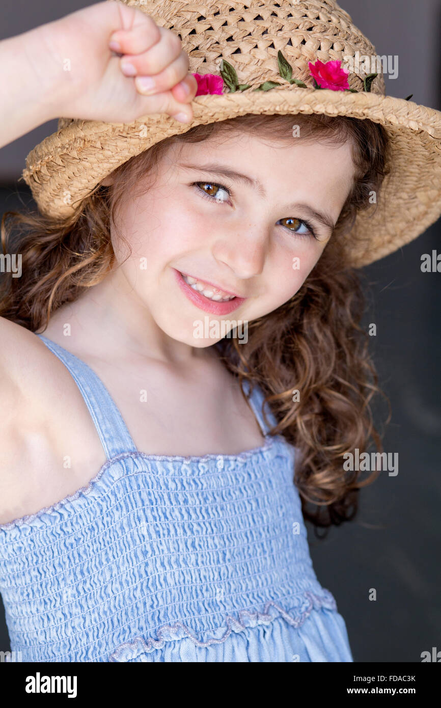 Portrait d'une jeune fille avec un fond gris. Elle porte une robe bleue et un chapeau de paille et souriant à la caméra. Banque D'Images
