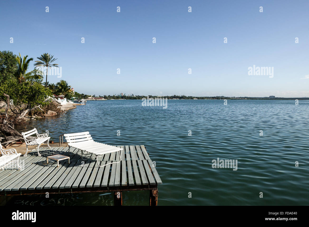 Terrasse en bois sur les pylônes au-dessus de la baie de Cienfuegos avec bancs blancs et chaise longue, ciel bleu au-dessus et rive bordée de palmiers., Cienfuegos, Cuba. Banque D'Images