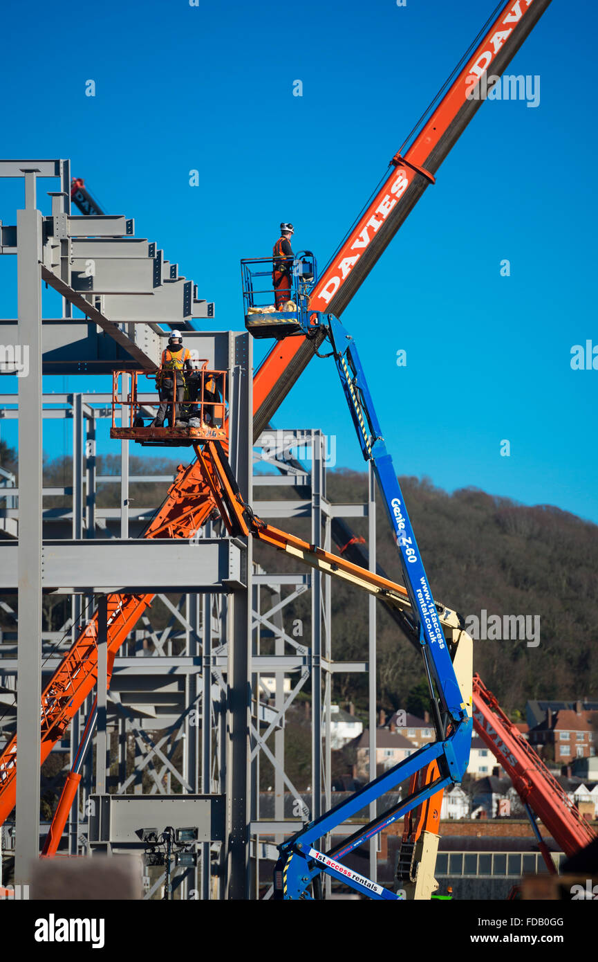 Les hommes travaillent en hauteur de 'grue' plates-formes élevées - construire ensemble le cadre en acier de boulonnage d'un nouveau bâtiment qui abritera une succursale de supermarché Tesco et Marks & Spencer store, sur un ciel bleu clair jour, Aberystwyth Wales UK Banque D'Images