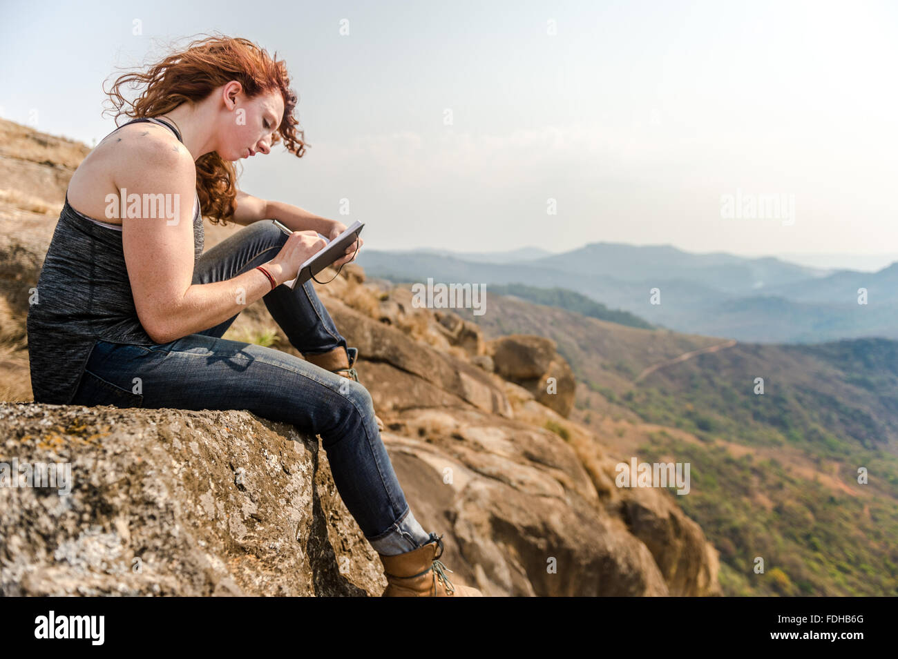 Fille à tête rouge écrit dans un journal en haut d'un rocher à Mlilwane Wildlife Sanctuary au Swaziland, Afrique. Banque D'Images