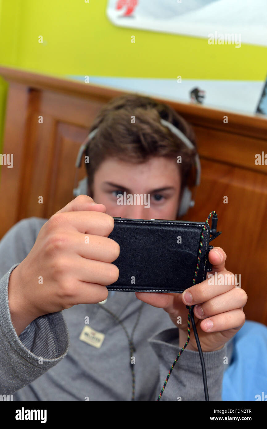Teenage boy se trouve sur son lit pendant l'utilisation de l'Internet sur son téléphone portable et écouteurs Parution Modèle Banque D'Images