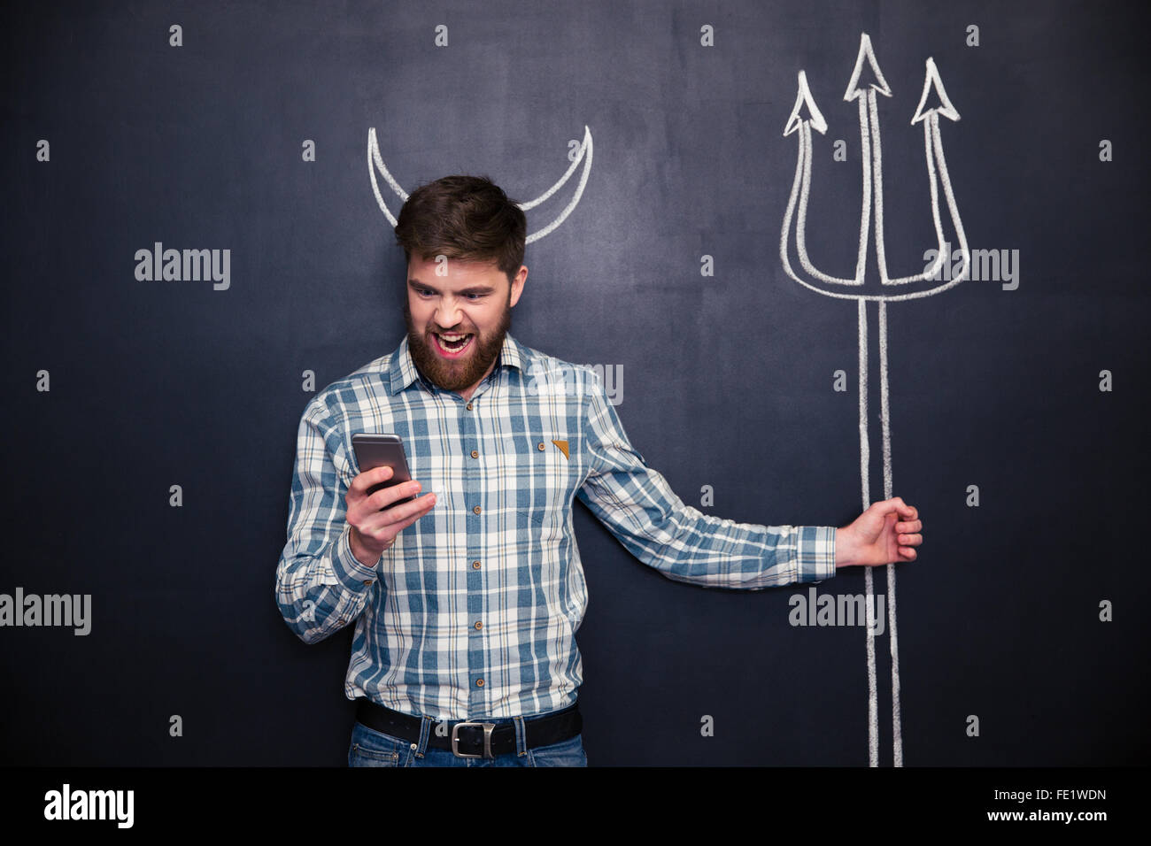 Beau jeune homme barbu jouer rôle de diable debout sur fond noir avec des cornes tirées et Trident Banque D'Images