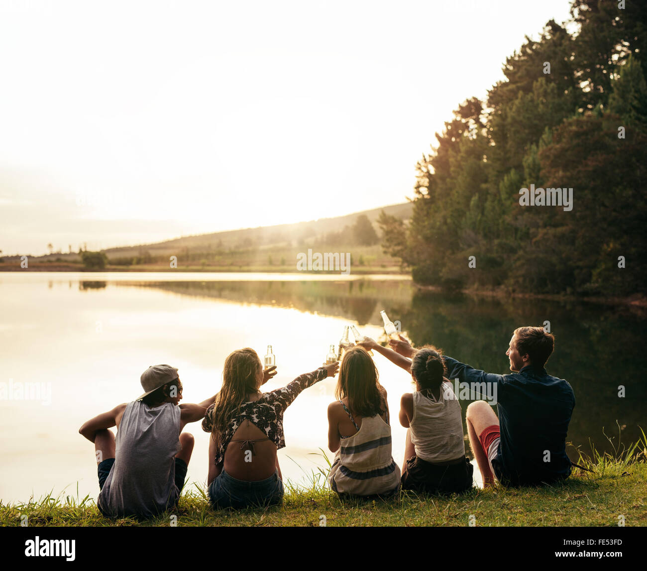 Groupe de jeunes gens assis dans une rangée à un lac. Young Friends toasting et célébrer avec bières au lac. Banque D'Images