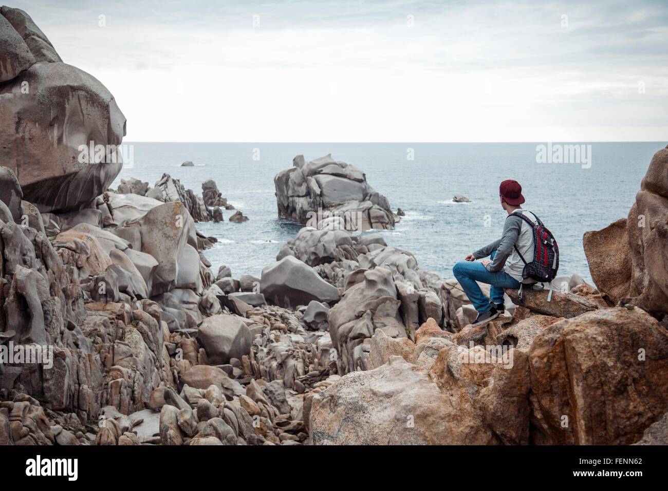 Vue arrière du jeune homme assis sur les rochers à la recherche en vue de l'océan, Costa Smeralda, Sardaigne, Italie Banque D'Images