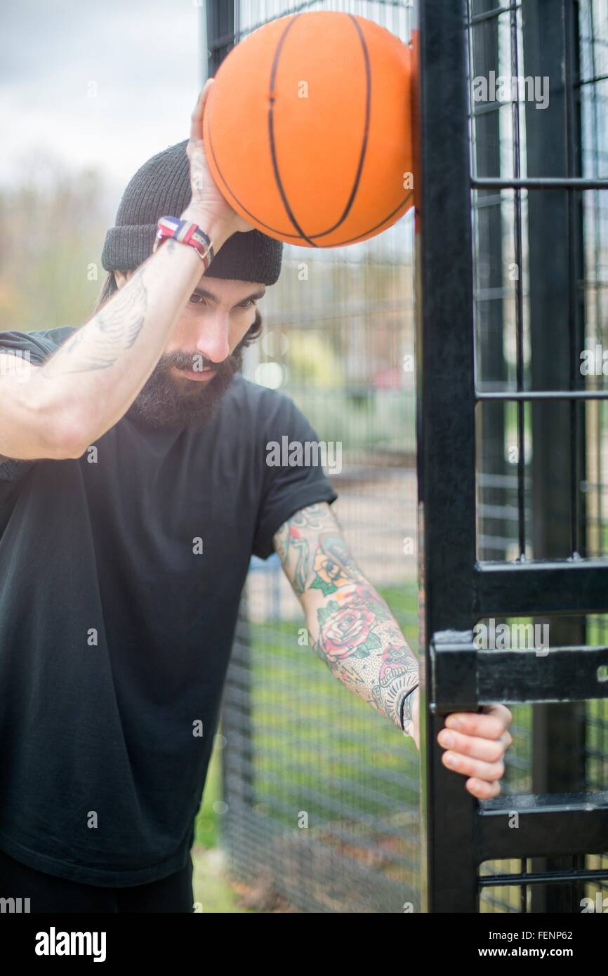 Mid adult man standing by fence, holding basketball contre barrière, pensive expression Banque D'Images