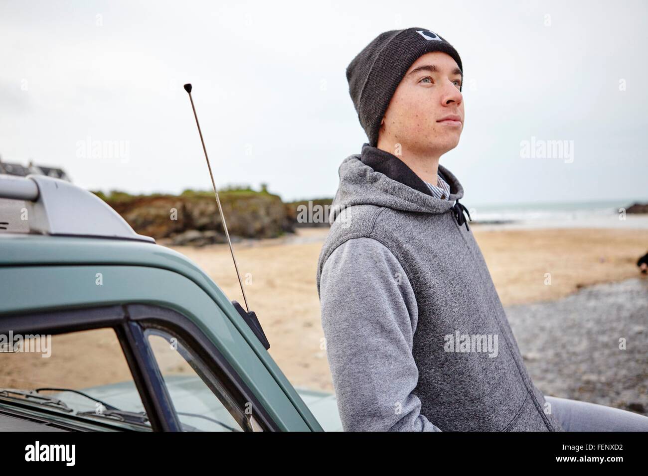 Young man leaning against car le regard vers le haut de la plage, à Constantine Bay, Cornwall, UK Banque D'Images