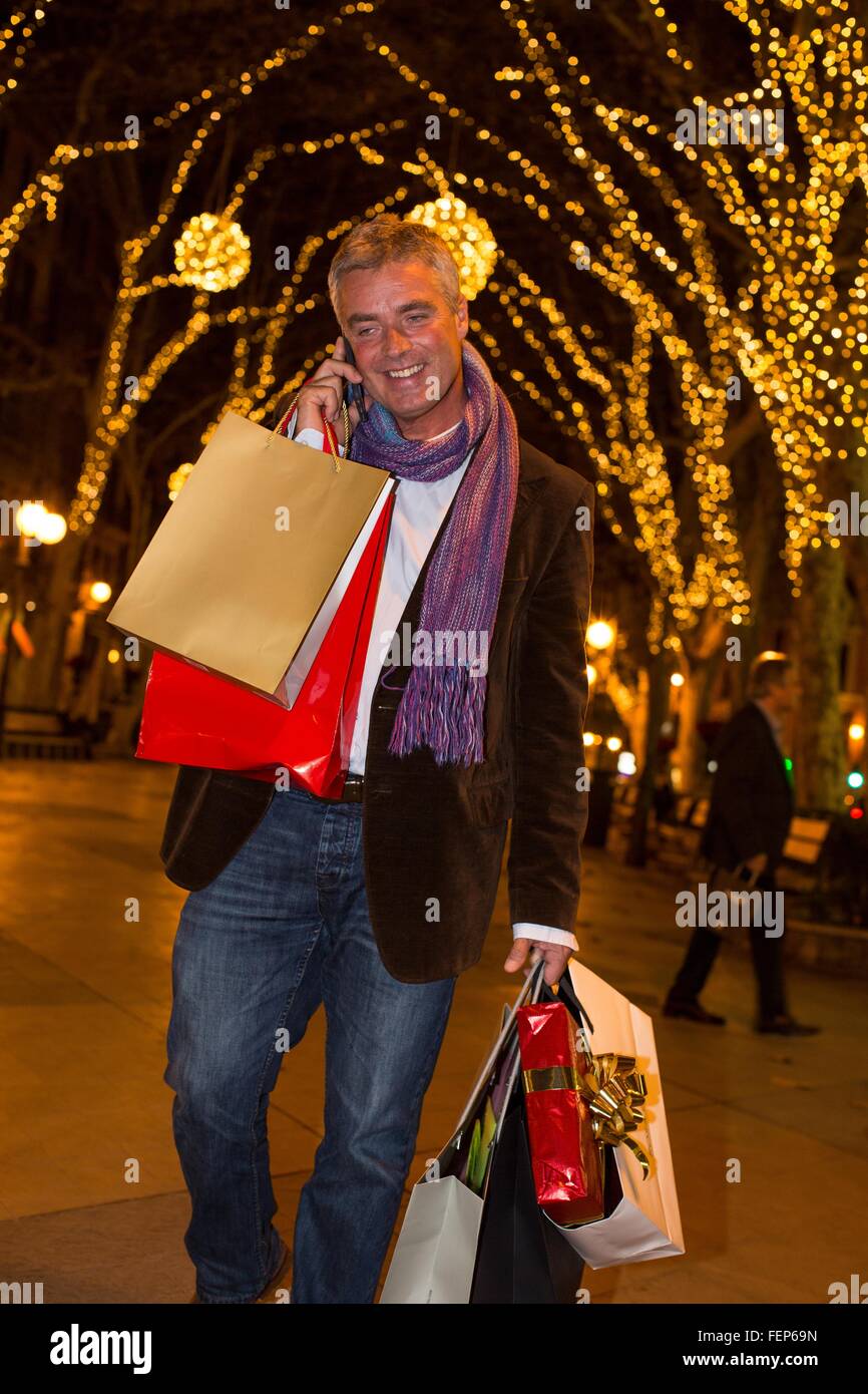 Homme mature avec xmas shopping talking on smartphone sur avenue ombragée d'arbres, Majorque, Espagne Banque D'Images