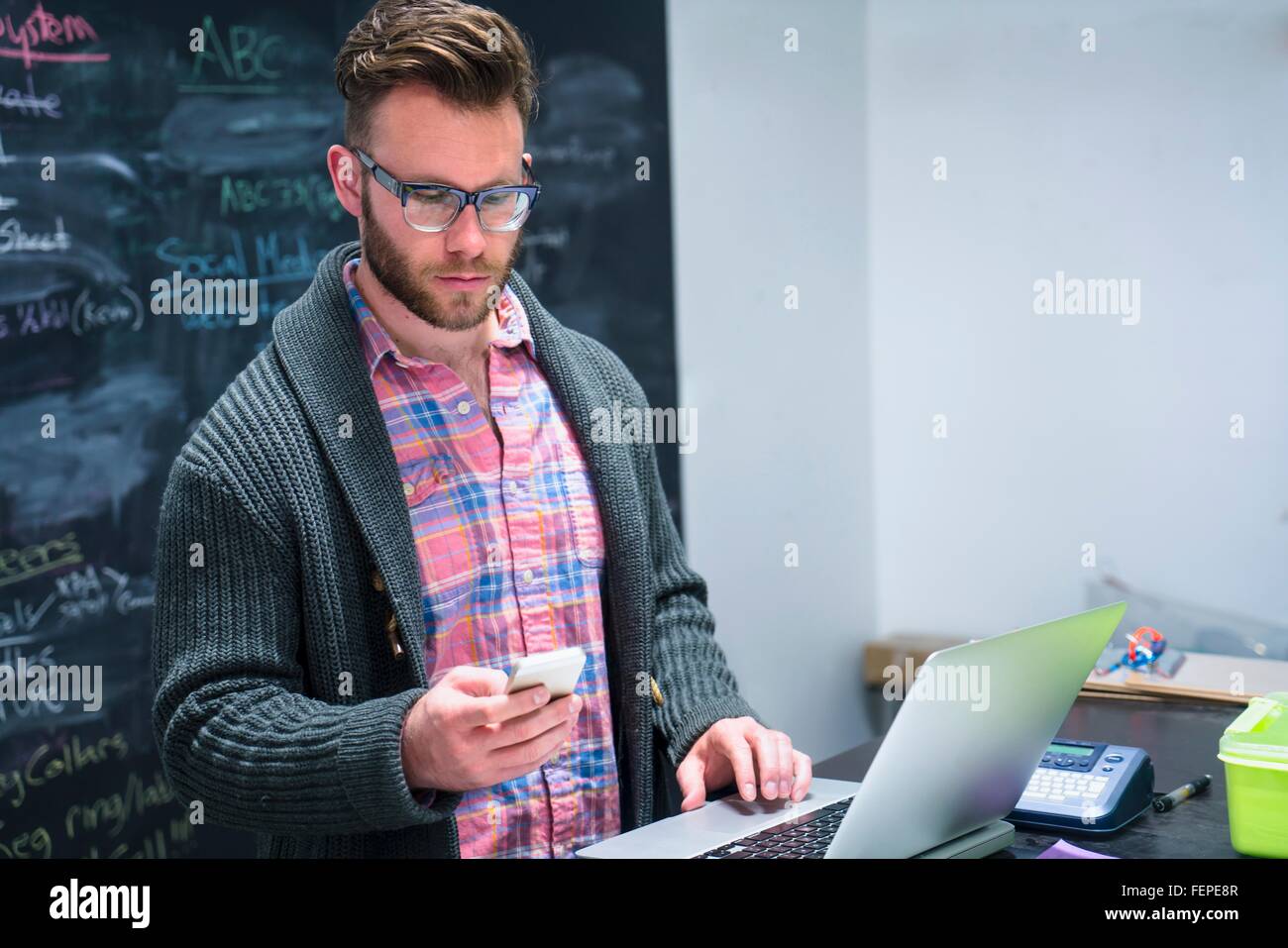 Jeune homme dans le lieu de travail à l'aide d'ordinateur portable looking at smartphone Banque D'Images