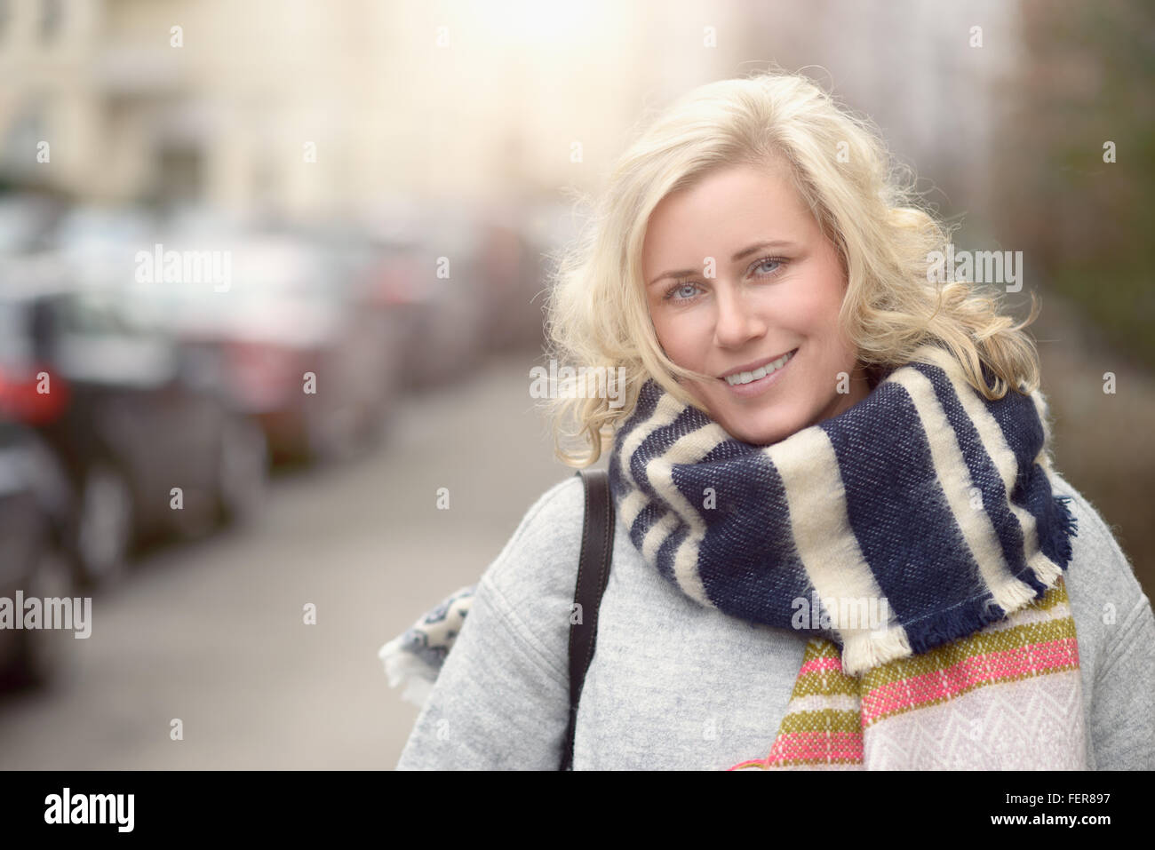 Smiling attractive young blonde woman dans une écharpe laineuse debout à l'autre d'une rue urbaine regardant la caméra, haut du corps Banque D'Images