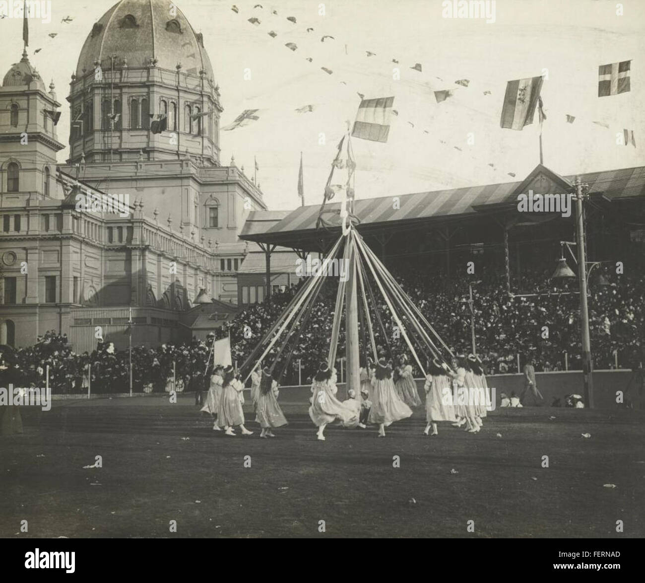 Une photographie historique d'une danse Maypole tenue devant l'école d'État, avec le bâtiment d'exposition en arrière-plan. La scène capture une célébration traditionnelle en plein air, mettant en vedette un événement communautaire animé. La danse Maypole, une activité symbolique, implique que les participants tissent des rubans colorés autour d'un poteau central. Le cadre souligne un moment important dans les rassemblements communautaires à un point de repère notable. Banque D'Images