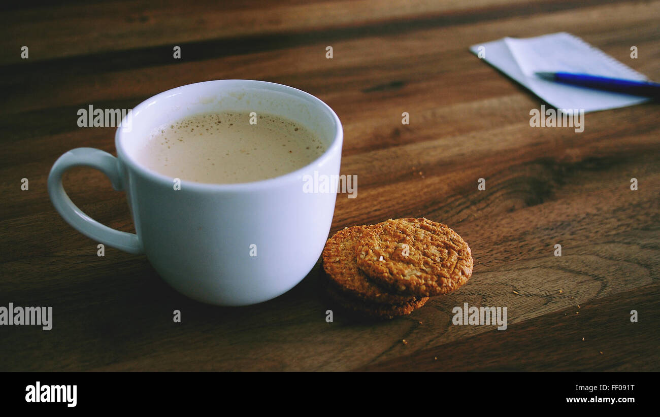 Un latte mousseux est assis à côté d'une assiette de biscuits, disposés dans une composition de nature morte. La tasse blanche contient la boisson crémeuse, créant une scène chaleureuse et accueillante. L'accent est mis sur la boisson chaude et réconfortante associée aux biscuits sucrés. Banque D'Images