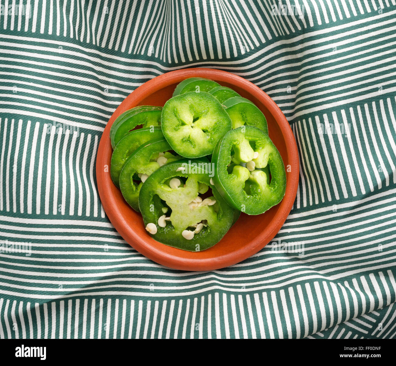 Vue de dessus d'un petit bol de piments jalapeño en tranches sur une nappe plissée vert et blanc éclairé avec lumière naturelle. Banque D'Images