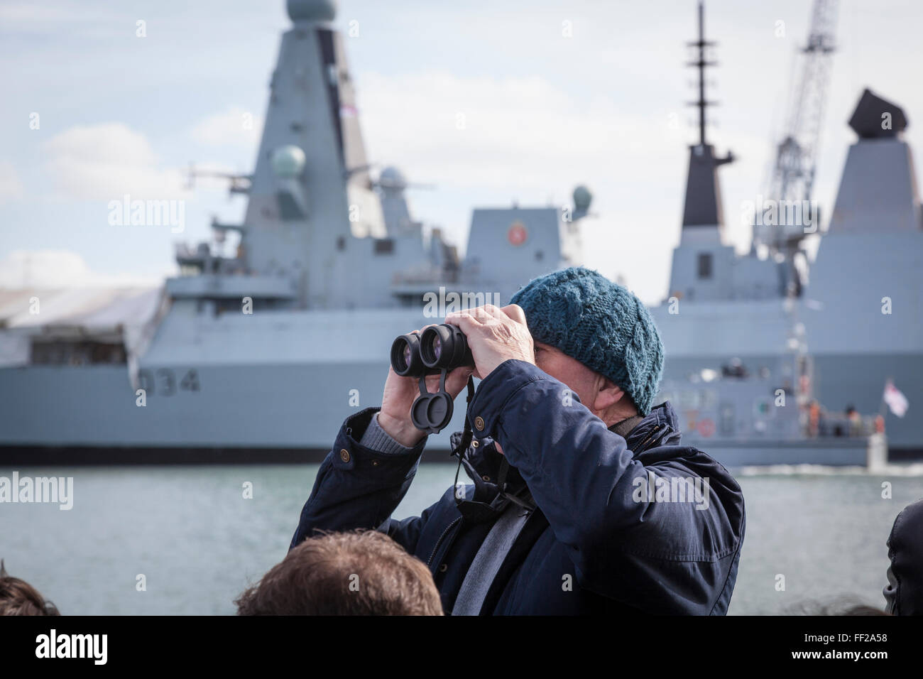 Un touriste sur une croisière dans le port de Portsmouth regarde à travers des jumelles alors que dans l'arrière-plan est accosté le HMS Diamond D34 un type 45 Banque D'Images