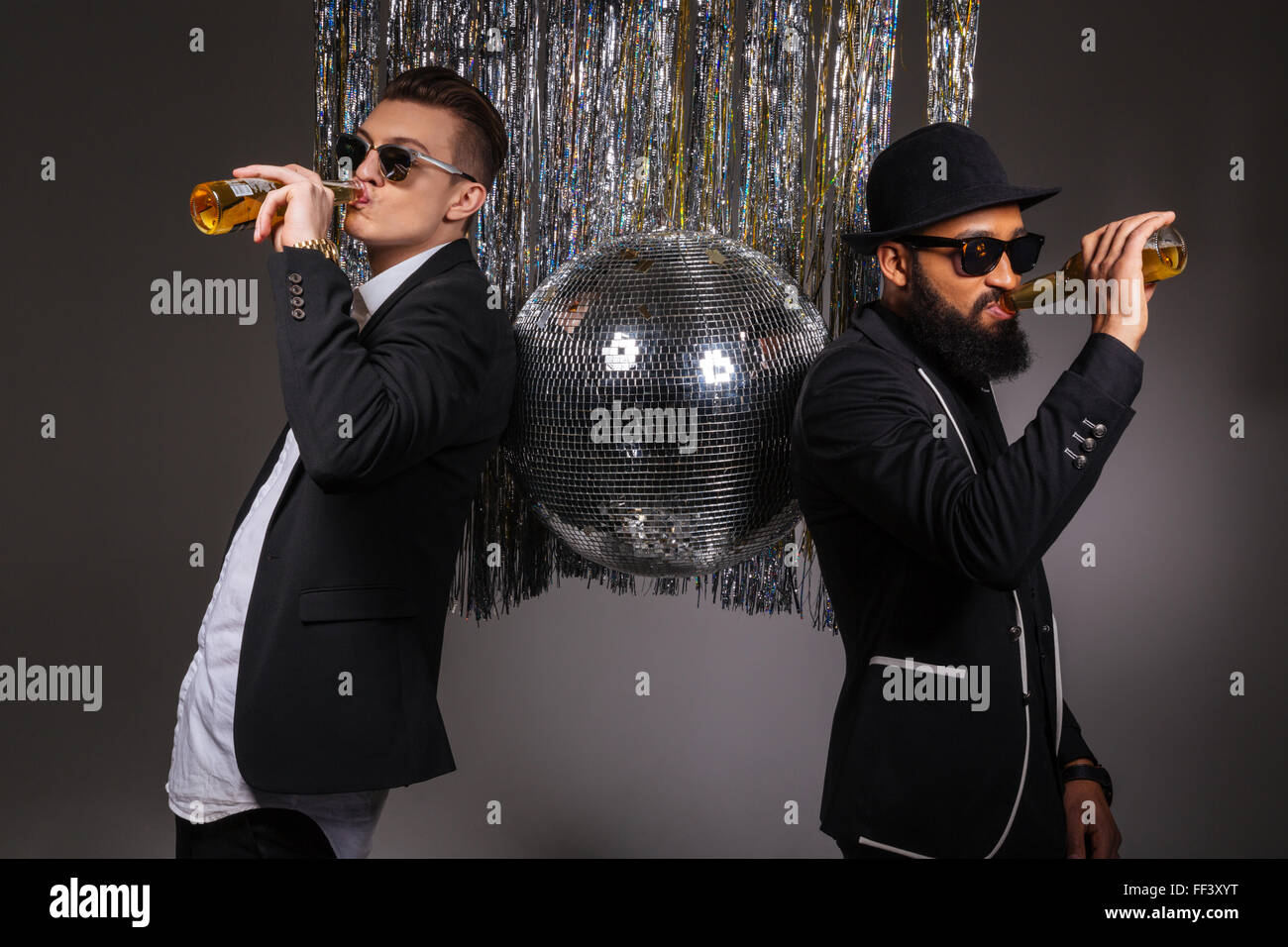 Deux beaux jeunes hommes confiants en costumes noirs et lunettes, debout près de boule disco et boire de la bière sur fond noir Banque D'Images