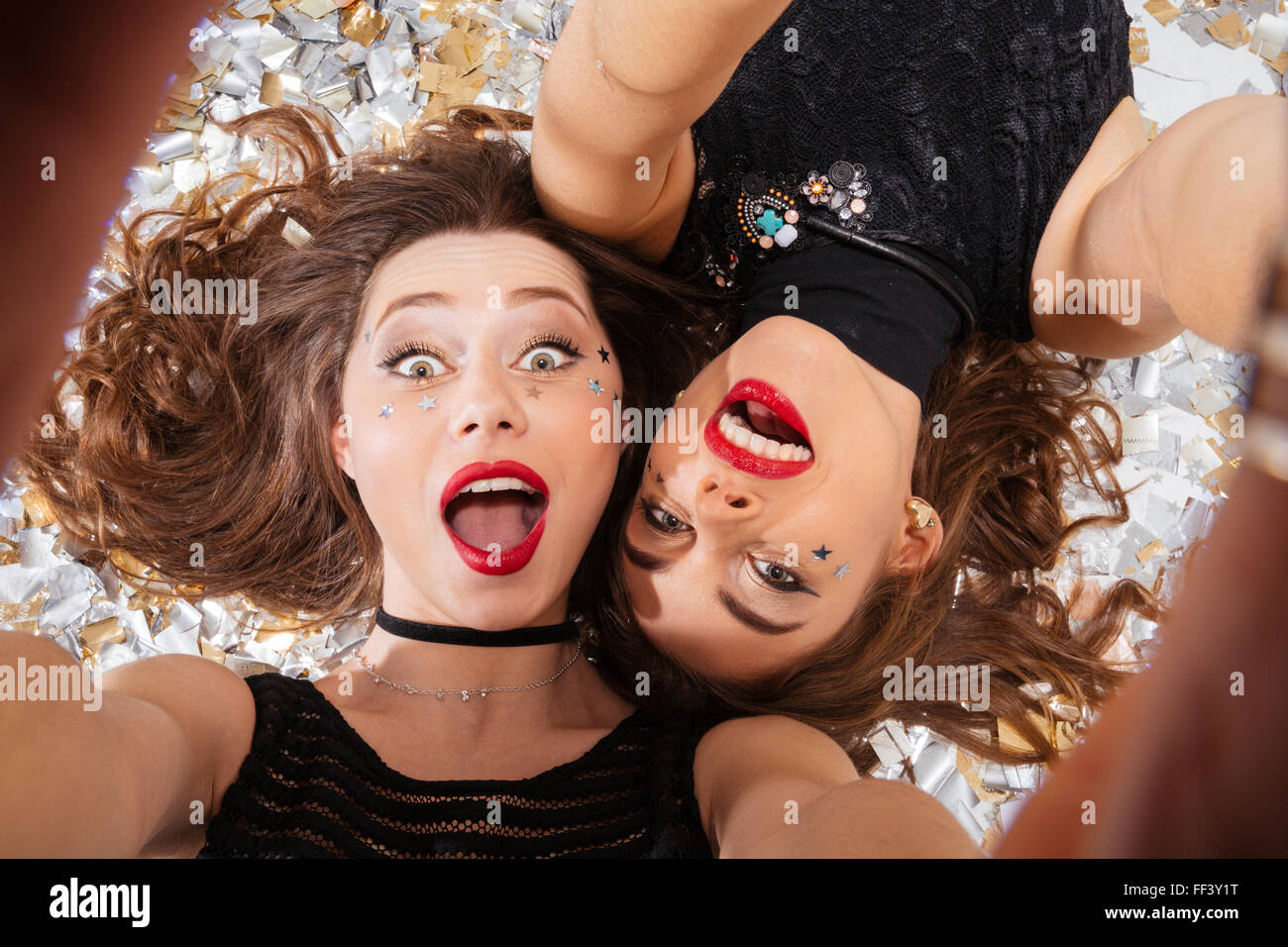 Deux jeunes femmes très excité avec lèvres rouges prenant et selfies couché sur fond de shining confetti Banque D'Images