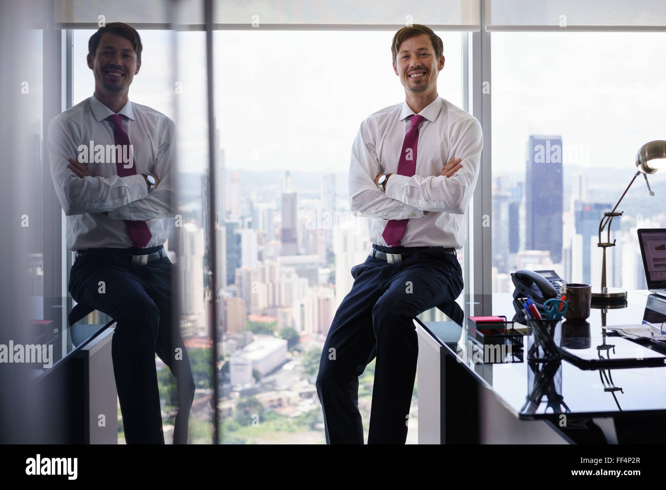 Adult businessman sitting in office moderne avec belle vue de la ville. L'homme regarde la caméra et sourit avec les bras cr Banque D'Images