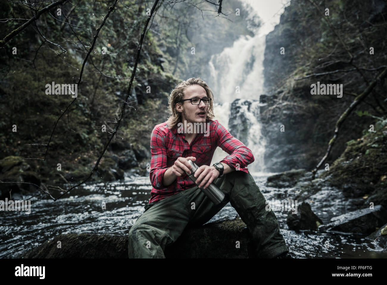 Un homme assis sur un rocher par une cascade, tenant une fiole. La randonnée à pied. Banque D'Images